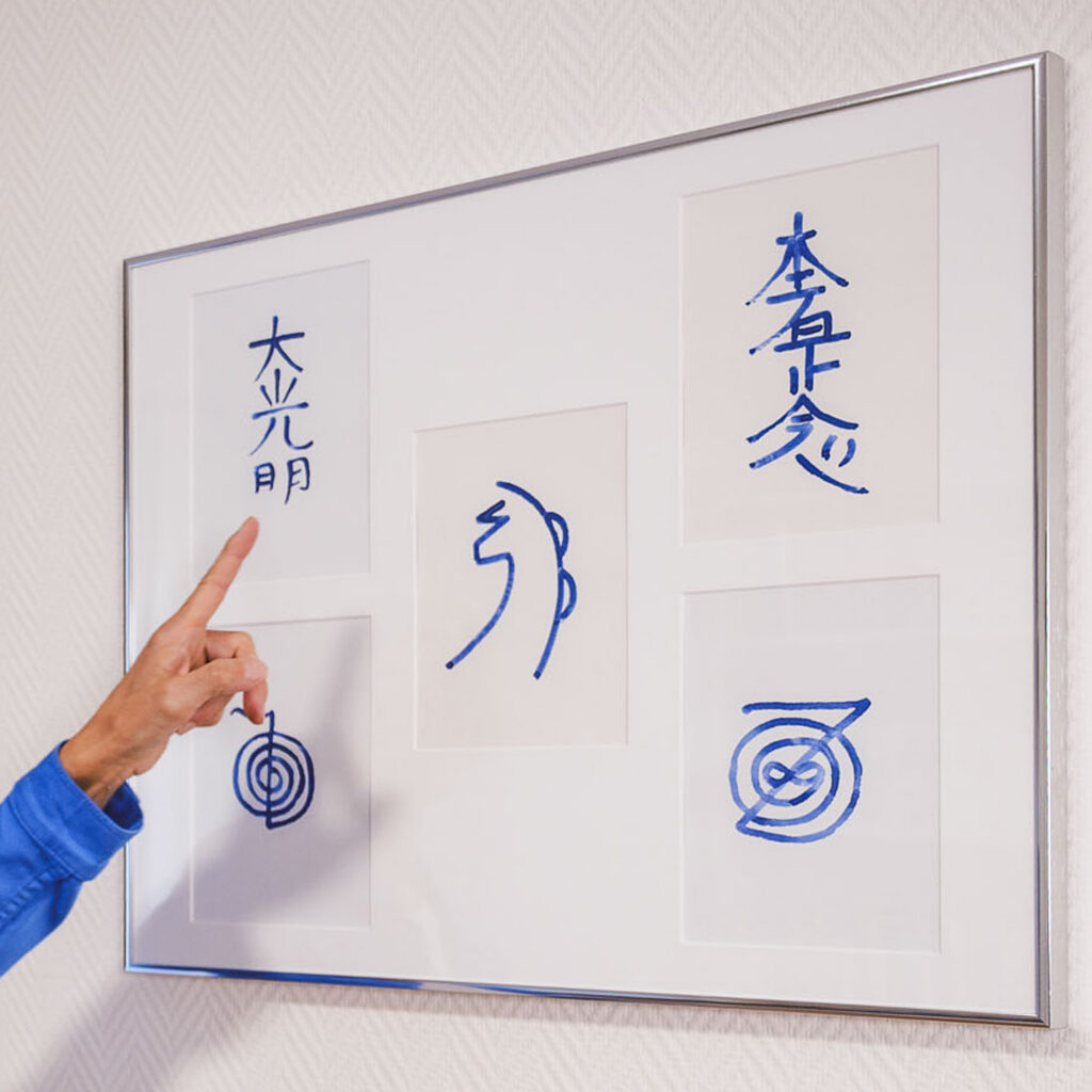 5 Hu-Man-Reiki© Symbole in blau auf weiß, in einem Bildrahmenan dr Wand. Eine Hand zeigt auf eines der Symbole.